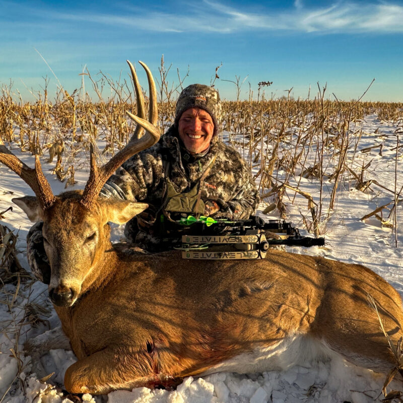John Dudley posing with a harvested whitetail deer in a snowy field, holding his compound bow, surrounded by harvested corn stalks under a clear blue sky.