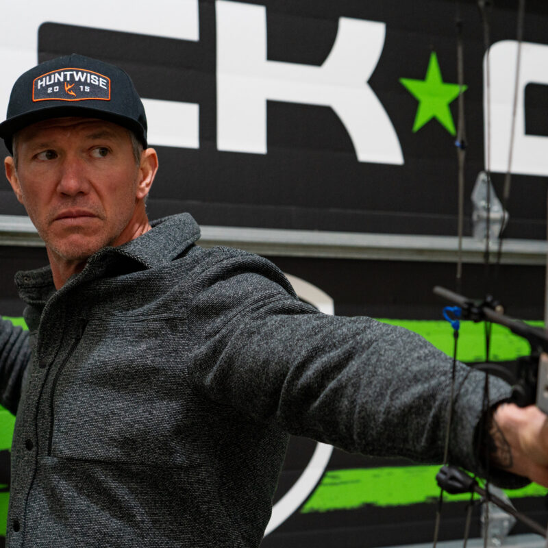 John Dudley, focused while drawing a compound bow in an indoor range with a branded background featuring bold green and black graphics.