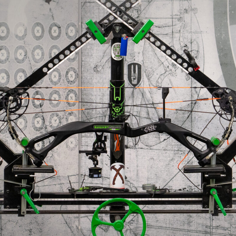 Compound bow mounted on a bow press, featuring green and black components, in a workshop setting with archery targets in the background.