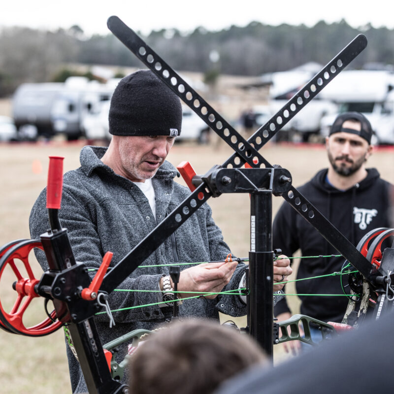 John Dudley working on a compound bow with a bow press at an outdoor event, with onlookers watching in the background.