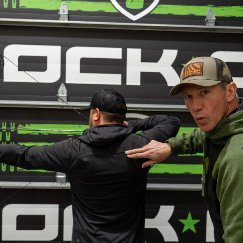 John Dudley instructing an archer on proper back tension technique in an indoor range, with a large ‘Nock On’ logo in the background.