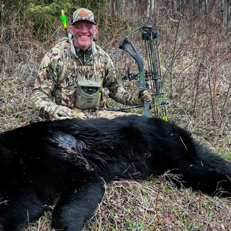 John Dudley smiling while kneeling next to a harvested black bear in a grassy, wooded area, holding his compound bow.