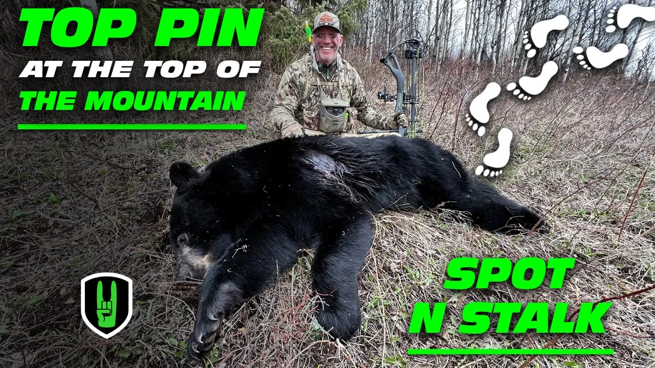 TOP PIN AT THE TOP OF THE MOUNTAIN - SPOT N STALK 1 John Dudley smiling while kneeling next to a harvested black bear in a grassy, wooded area, holding his compound bow.
