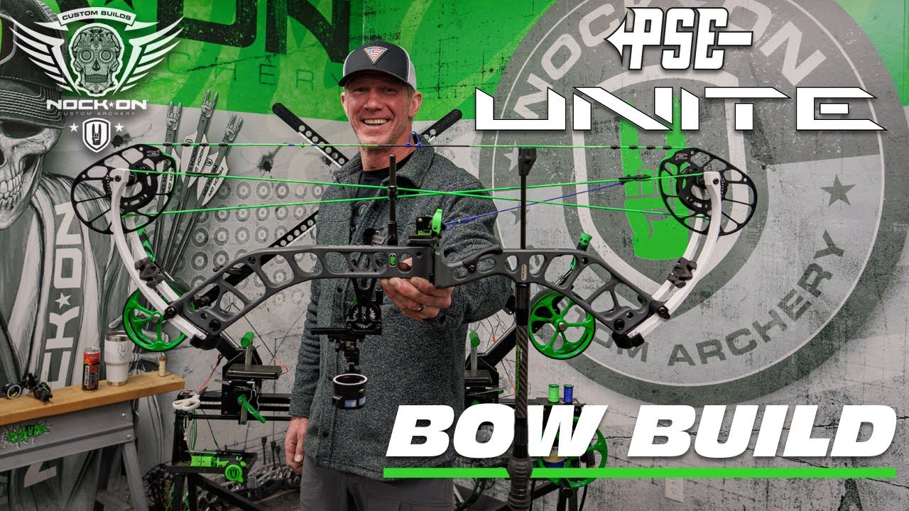 John Dudley smiling while holding a compound bow in the Nock On Archery workshop, surrounded by archery equipment with the words bow build.