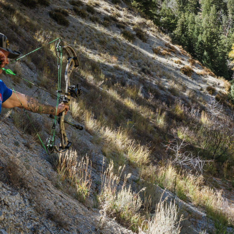 John Dudley aiming a compound bow on a steep, rocky hillside, with trees and vegetation in the background during an outdoor practice session.