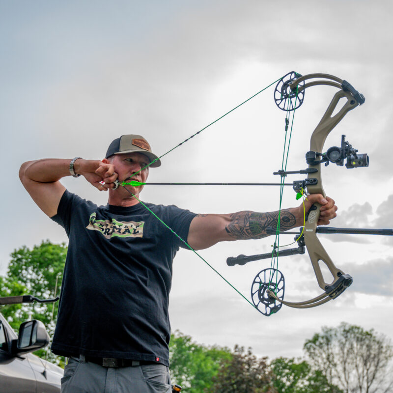 John Dudley drawing a compound bow outdoors, aiming with focus, while standing next to a vehicle and archery equipment, under a cloudy sky.