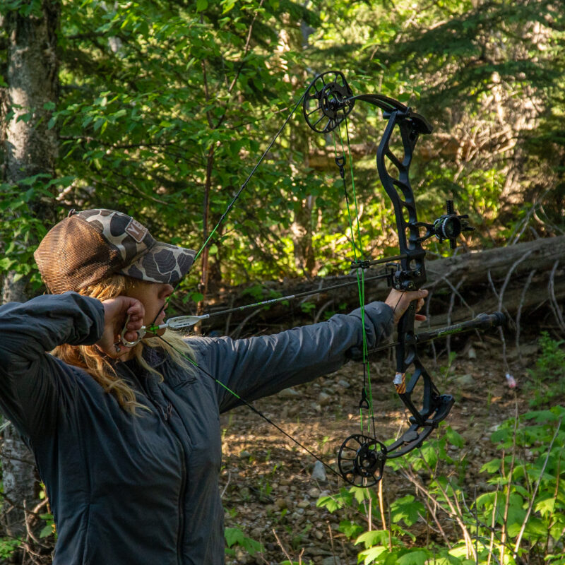 Woman at full draw in the woods with a compound bow.