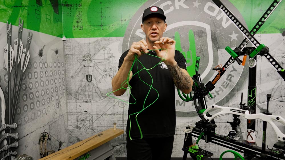 Bow Specs Not Matching Up? Post-String-Change Adjustments Made Easy 7 John Dudley holding a bowstring while discussing its characteristics, with archery equipment and instructional materials visible in the background.