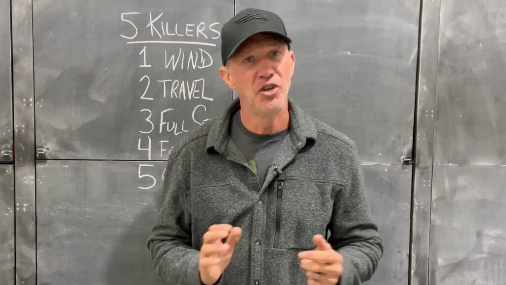 School Of Nock 6 A man wearing a dark gray jacket and black cap is speaking in front of a chalkboard.