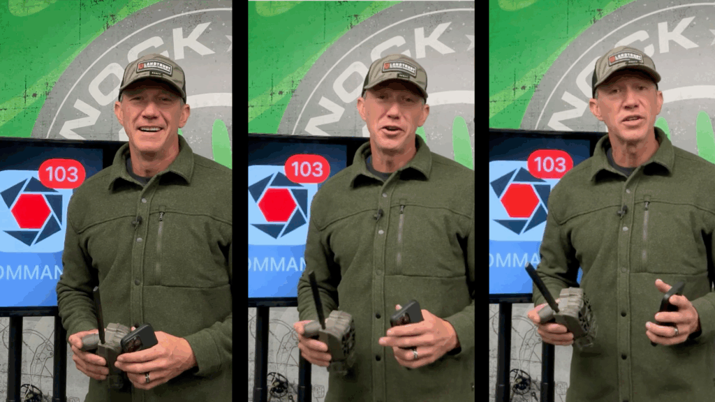 School Of Nock 3 A smiling man wearing a green jacket and a baseball cap stands indoors holding a trail camera and a smartphone.