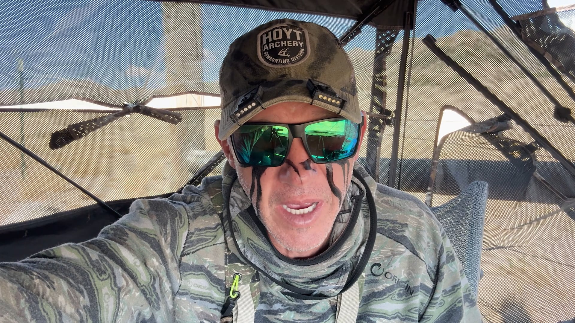 Person in camouflage gear and Hoyt Archery cap wearing reflective sunglasses.