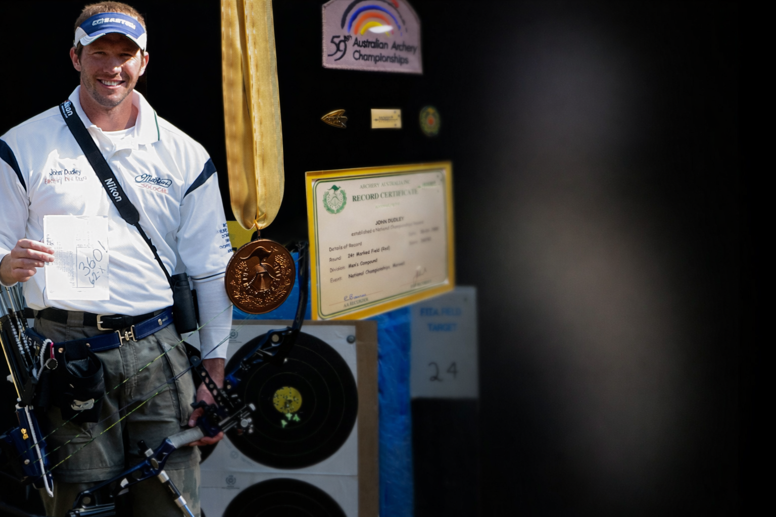 A man holding a compound bow and a scorecard stands next to archery targets.