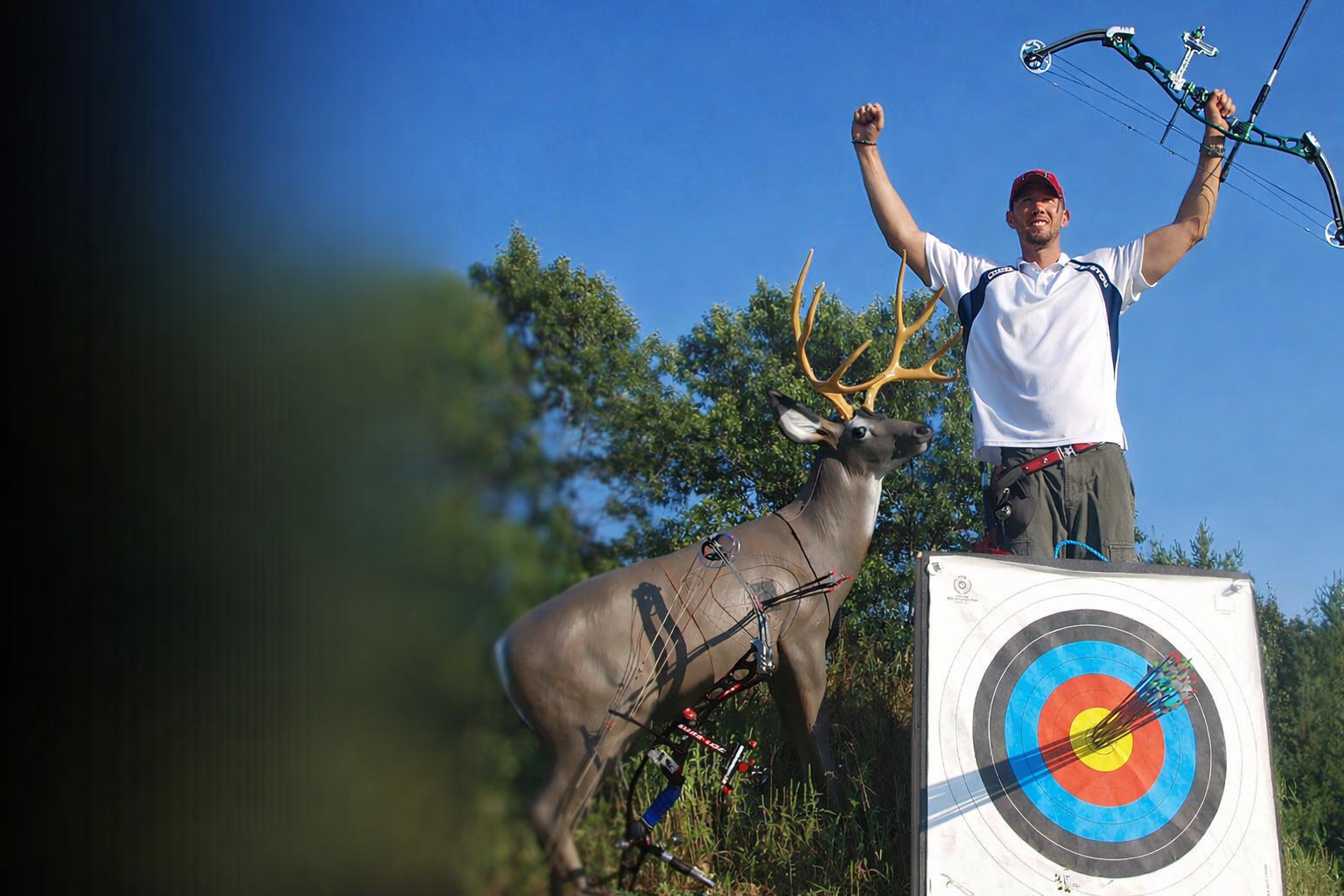 Archer celebrates with compound bow raised, standing behind a deer target and a bullseye target with arrows grouped in the center.