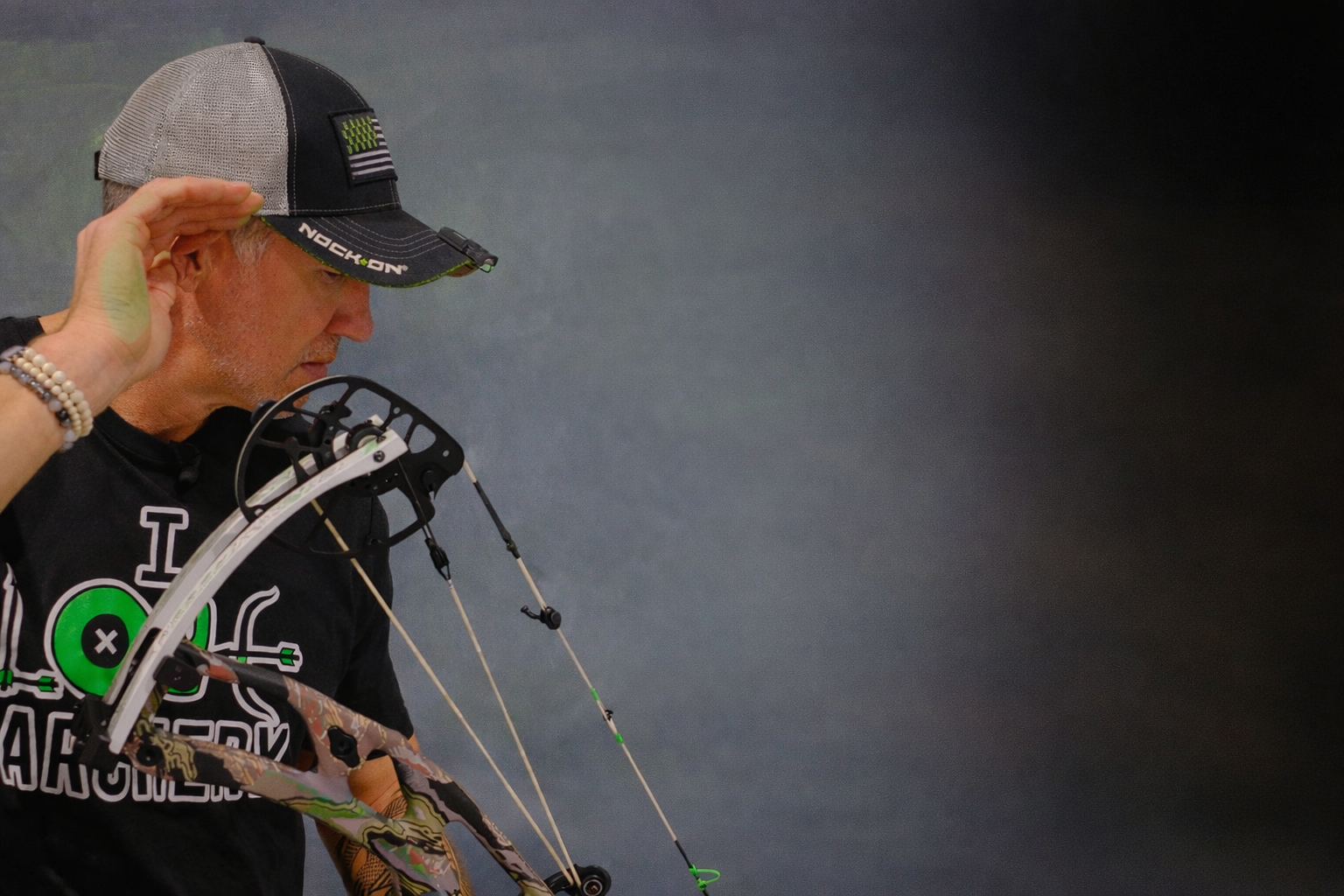 A man in a black Nock On archery t-shirt and gray trucker cap holds a compound bow while touching the brim of his hat