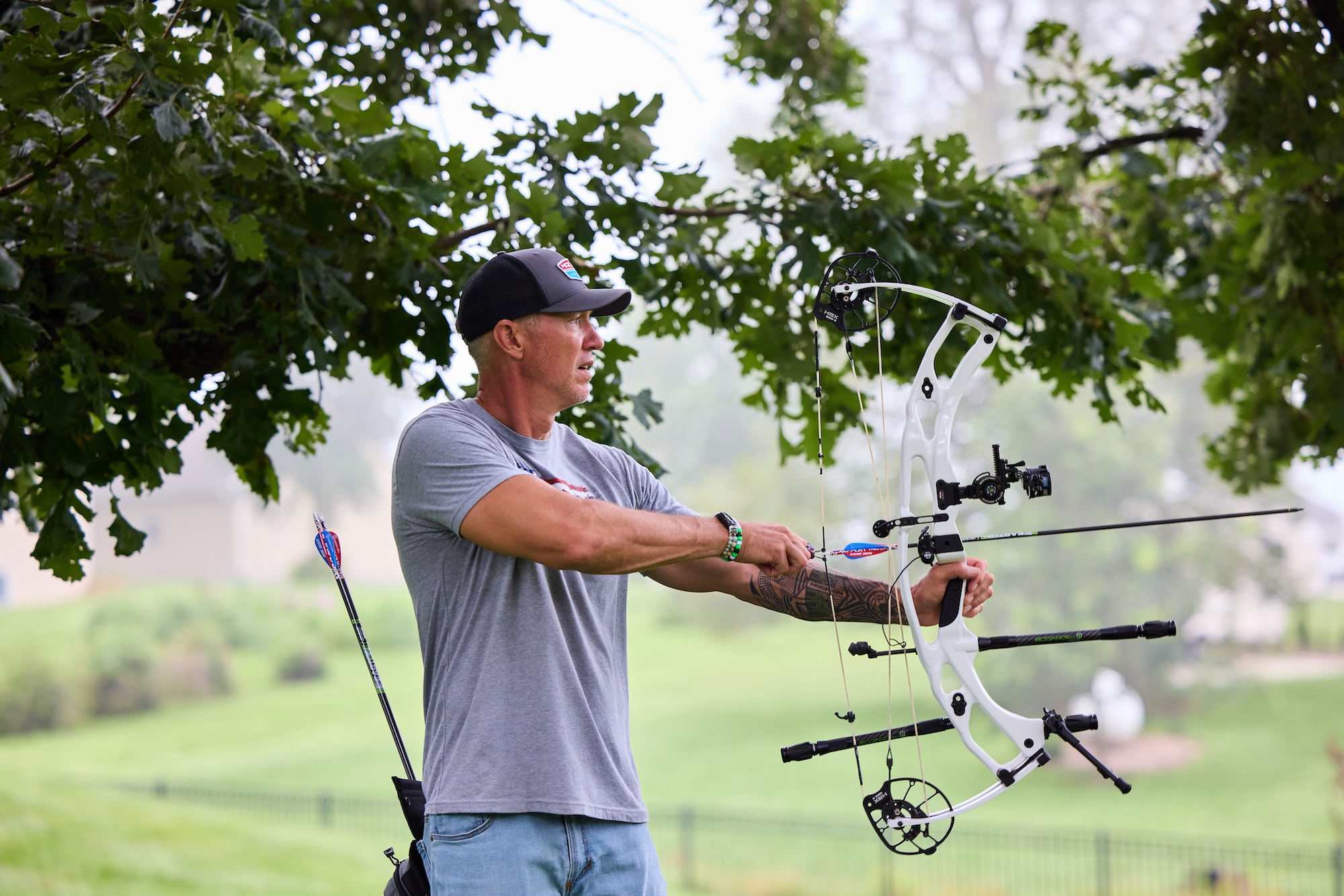 Archer draws a white compound bow while aiming outdoors on a shaded green lawn.